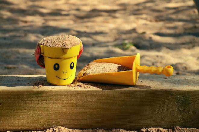 Sand inside a yellow bucket with a smiley face and a yellow scoop on the wooden edge of a sand pit